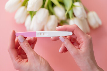 Positive pregnancy test in a woman's hand on a pastel pink background.