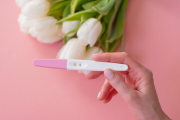 Positive pregnancy test in a woman's hand on a pastel pink background.