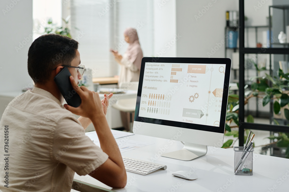 Poster Young busy white collar worker with mobile phone by ear sitting by workplace and looking at computer screen with data