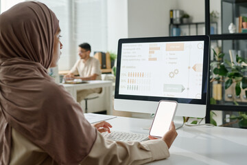 Young Muslim businesswoman in hijab holding smartphone with blank screen while sitting by workplace in front of desktop computer
