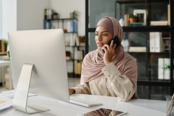 Young confident Muslim businesswoman in hijab looking at computer screen and speaking on mobile...