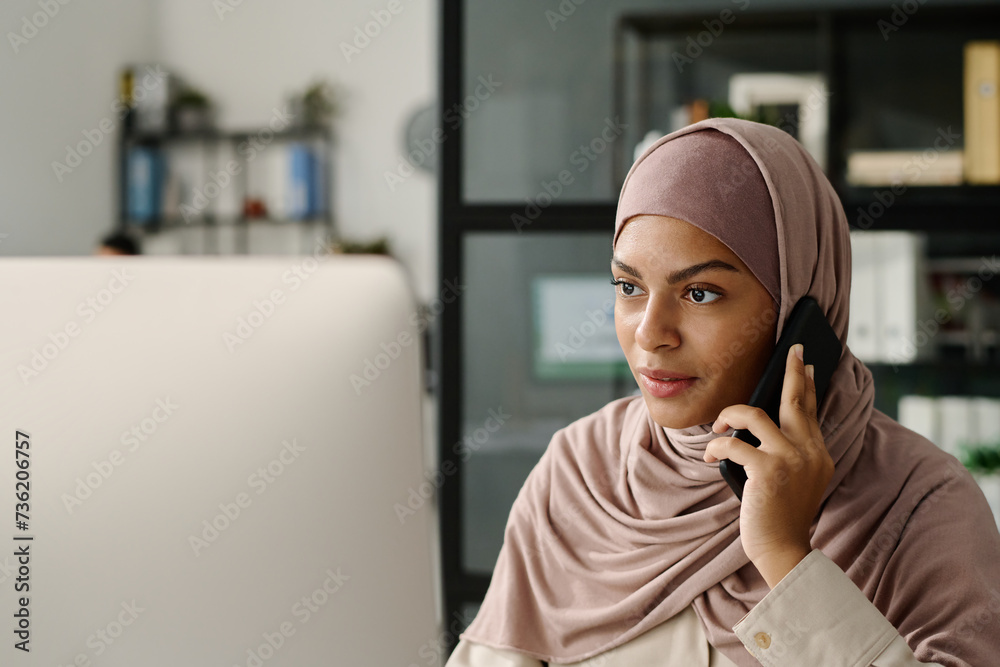 Poster confident muslim white collar worker in hijab talking to applicant on mobile phone and looking at co