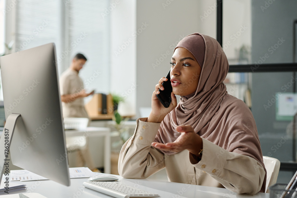 Poster young muslim female employee in hijab explaining something to business partner on phone while sittin