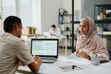 Two young intercultural Muslim business people looking at one another at meeting by workplace during discussion of working points