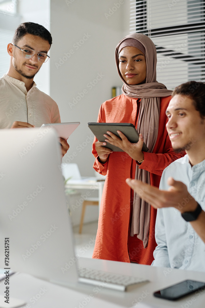 Poster two young muslim brokers with tablets looking at computer screen while listening to male colleague e