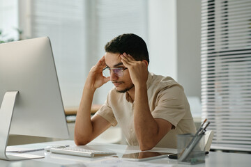 Young stressed male employee sitting by desk in front of computer screen and waiting for page uploading while trying to concentrate