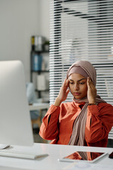 Young tired female manager touching her head while sitting by workplace with desktop computer and suffering from headache