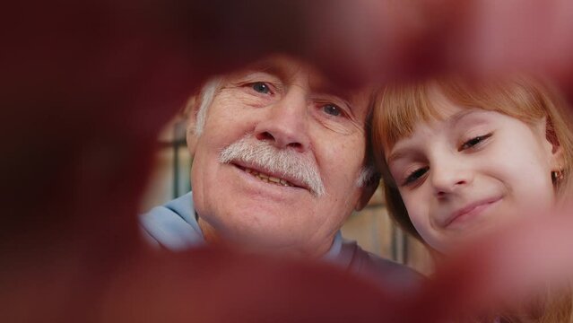 I love you. Happy Caucasian grandfather and granddaughter making symbol of love, showing heart sign to camera, express feelings sincere positive feelings. Cute family sitting on sofa in living room.