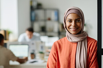 Happy young Muslim female entrepreneur or manager in hijab looking at camera with smile while standing in coworking space