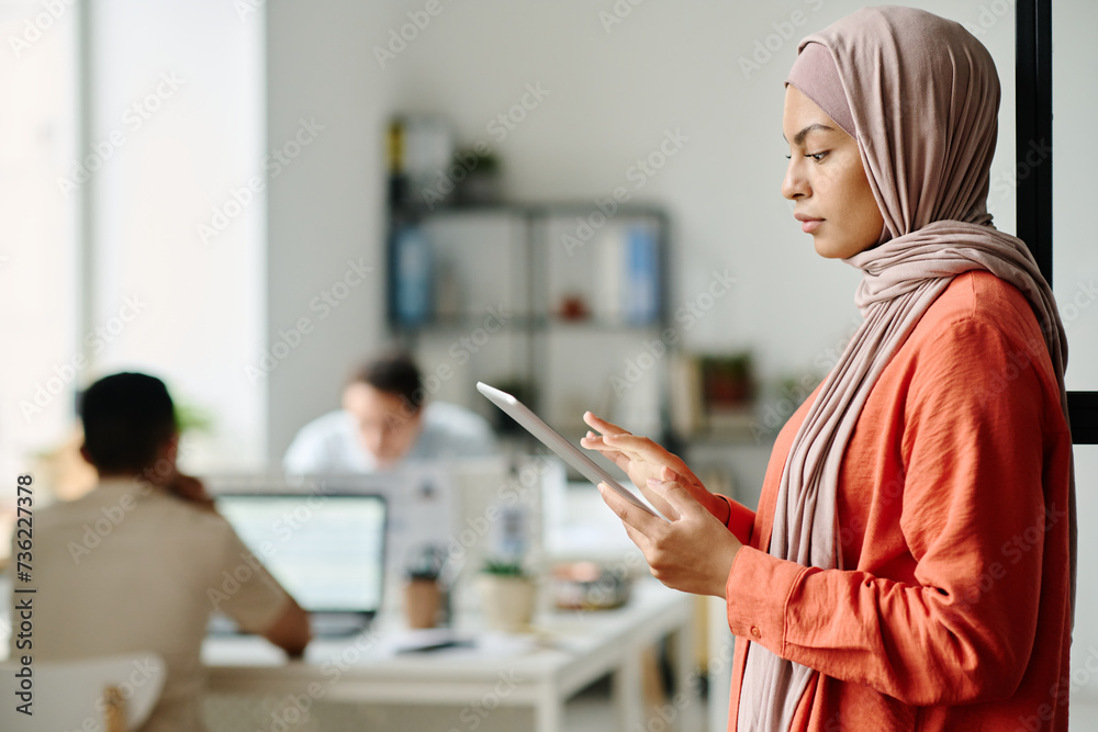 Aufkleber young muslim female employee in hijab using tablet while standing in front of camera in coworking sp