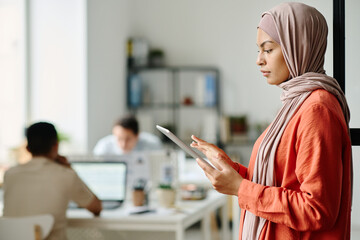 Young Muslim female employee in hijab using tablet while standing in front of camera in coworking...