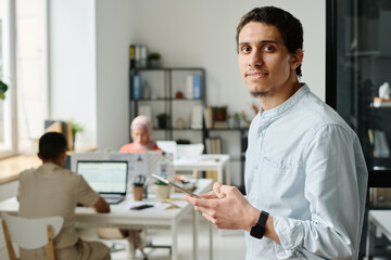 Naklejka na ściany i meble Young Muslim male entrepreneur or office manager with tablet looking at camera with smile while standing against colleagues in coworking space