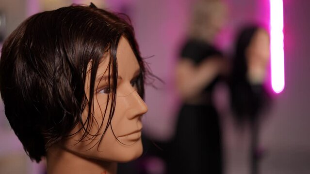Close-up. Mannequin of a male head in a short wig. In the background, out of focus, a girl is combing a mannequin of a female head with long hair with a comb