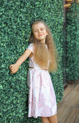 Cute blond Caucasian girl with a smile in a pink dress. Background - Green living wall. Happy childhood concept