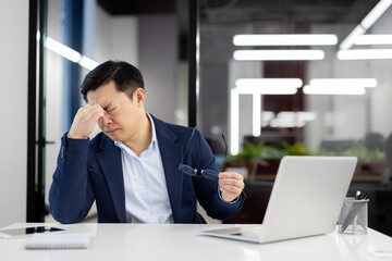 Overworked and overtired man has severe dizziness in head, businessman rubs eyes, eye pain long work at laptop, asian man inside office at workplace.