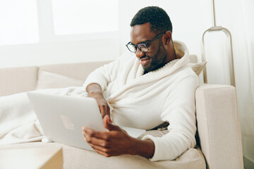 Smiling African American Man in Bathrobe Working on Laptop in Modern Living Room The young freelancer is sitting on the sofa, typing on his computer in the morning He exudes a happy and relaxed vibe