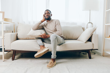 Relaxed African American man sitting on a comfortable sofa in his modern living room, smiling while enjoying a peaceful weekend at home He holds a pillow and appears to be deep in thought, perhaps
