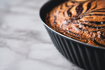 Zebra cake with a striped pattern baked in a metal dish, placed on a white marble tabletop, close-up view photo with copy space.