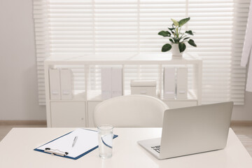 Doctor's workplace. Laptop, clipboard and glass of water on white table in hospital