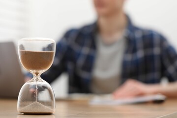 Hourglass with flowing sand on desk. Man taking notes while using laptop indoors, selective focus