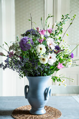 Blumenstrauss in einer Vase aus Keramik vor einer Glastür. Frühlingsblumen: Kamille, Eustoma, Anemone, Spirea, Allium, Strandflieder, Zweige