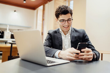 Smiling young businessman in a suit using smartphone and laptop at a cafe, embodying multitasking and modern work lifestyle.