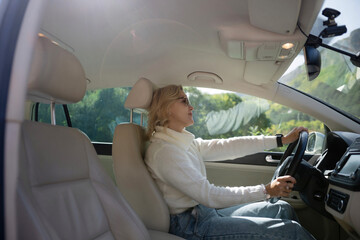 A blonde woman in a white sweater and jeans is driving. Happy woman sitting in a car with a white interior.