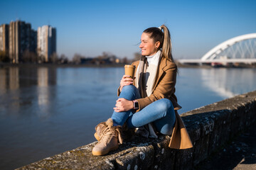 Fototapeta na wymiar Beautiful woman in warm clothing enjoys drinking coffee and resting by the river on a sunny winter day. 