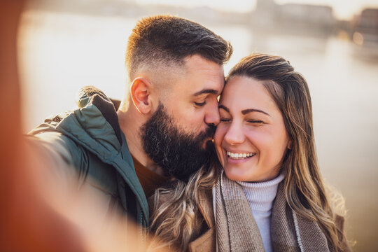 Close up image of happy couple taking selfie outdoor. Man kissing his woman in cheek.