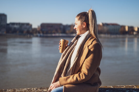Beautiful woman in warm clothing enjoys drinking coffee and resting by the river on a sunny winter day. Toned image.	