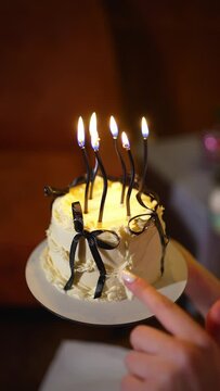 Vanilla birthday cake with lit candles and dark ribbons. Close-up image of person hand eating cream from holiday tsake. Festive moments