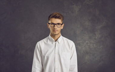 Fototapeta premium Studio portrait of serious young businessman or programmer standing on gray background. Skinny Caucasian man wearing white shirt and glasses looks straight into camera. Confidence concept.