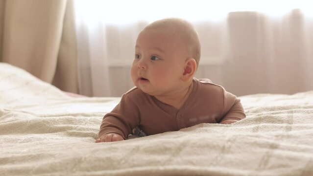 the baby lies on his stomach at home on the bed and looks around the baby is exploring the world close up in slow motion