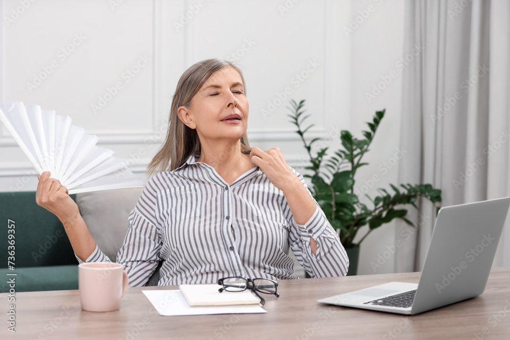 Sticker Menopause. Woman waving hand fan to cool herself during hot flash at wooden table indoors