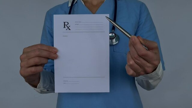 A female doctor in a blue uniform is holding a blank prescription pad and pen. She is looking at the camera with a serious expression