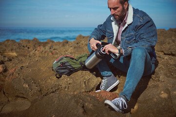 Confident portrait of a mid adult Caucasian bearded traveler man, hiker adventurer opening the stainless steel thermos flask, enjoying happy day alone on the nature, sitting by sea on the rocky cliff