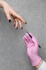Hand of a nail technician in pink gloves with a brush and a client's hand against gray background.