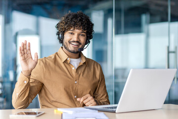 Portrait of a smiling Indian young man wearing a suit sitting in the office at a desk with a laptop, looking and waving at the camera.