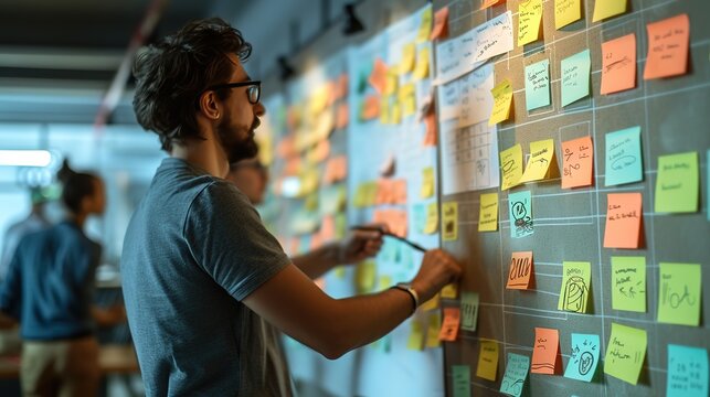 Focused young male professional organizing ideas using colorful sticky notes on a glass wall in a collaborative workspace.