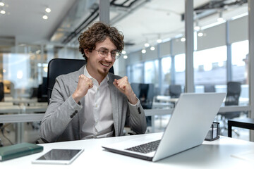 A young entrepreneur with curly hair shows excitement in the office, manifesting success at his workplace with a laptop in front.