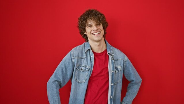 Cheerful young man with curly hair smiling in denim jacket against a vibrant red background