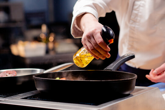 close up chef in a white uniform sprays a frying pan with oil from a spray bottle in professional kitchen