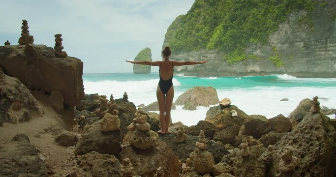 Woman in black swimsuit stands with outstretched arms among the stone cairns at Tembeling Beach, Nusa Penida. Ocean waves crash behind her.