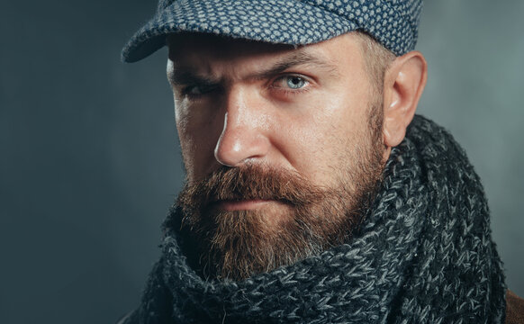 Closeup portrait of serious man with beard in cap and scarf. Winter accessories. Handsome bearded man in gray scarf and hat. Attractive male in fashionable clothing. Autumn-winter fashion for men.