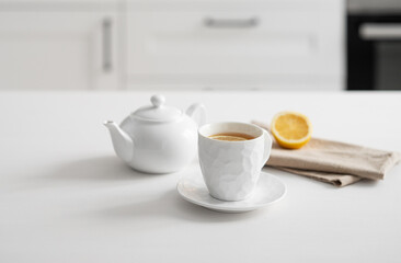 A cup of tea with lemon and a teapot on a white table against the background of a white kitchen.
