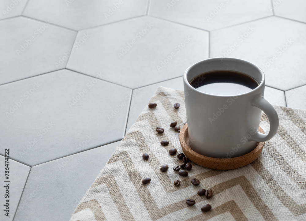 Wall mural A cup of black coffee on a napkin on a trendy gray tile background with coffee beans and morning light from the window.