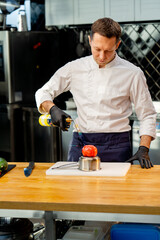 the chef is standing in the kitchen in a white jacket holding torch and about to burn a tomato