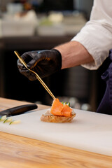 close-up of hands in black gloves placing piece of salmon on bread with golden tweezers