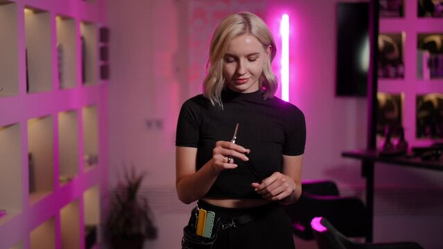 Beautiful girl barber posing while standing in a fashionable barbershop. The girl looks at the hairdressing scissors and a comb in her hands. The girl looks at the camera. Slow motion