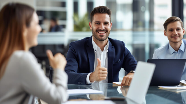 A confident professional man in a suit is giving a thumbs up in a bright office environment with cheerful colleagues around him, indicating success and approval.